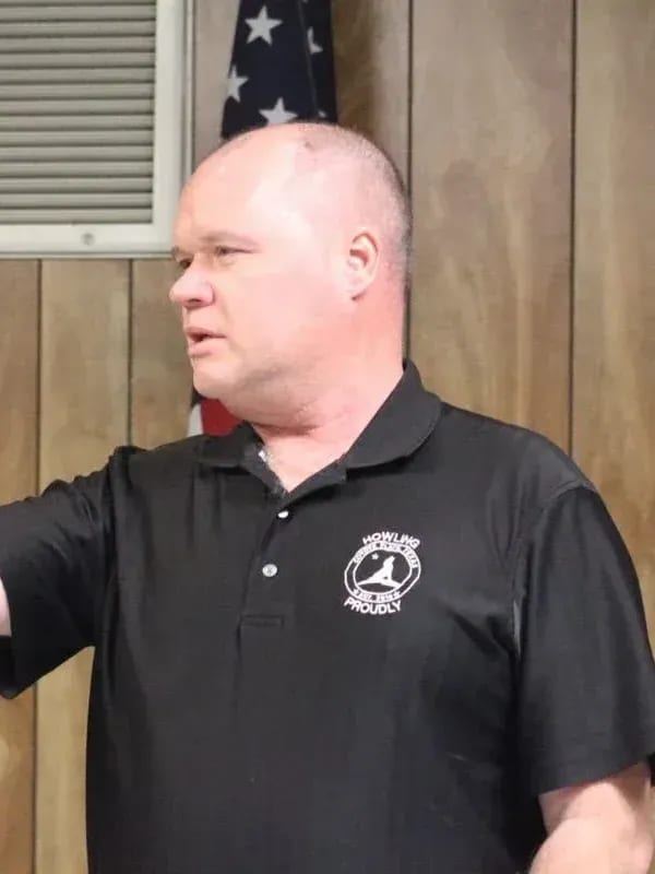 A man in a black shirt is standing in front of an american flag